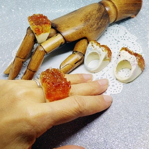 May include: Four rings with a unique design. The rings are made of white ceramic with a textured surface and feature a colorful, translucent, resin-like material. The rings are displayed on a wooden hand and a white doily.