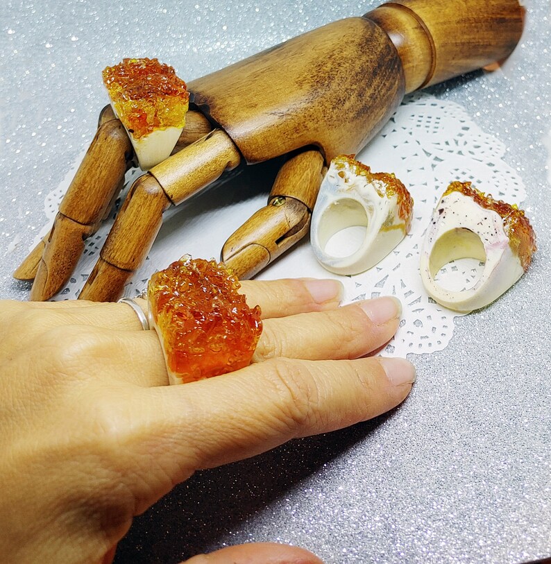 May include: Four rings with a unique design. The rings are made of white ceramic with a textured surface and feature a colorful, translucent, resin-like material. The rings are displayed on a wooden hand and a white doily.