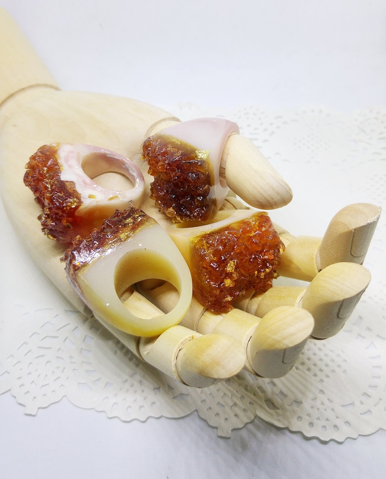 May include: Four resin rings with a white base and a brown, glittery top. The rings are displayed on a wooden hand.