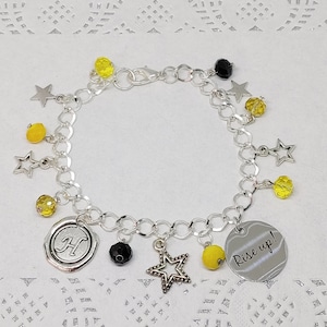 Peut inclure: Un bracelet à breloques en argent avec des perles jaunes et noires, des breloques en forme d'étoile et deux breloques avec la lettre "H" et les mots "Rise up!"