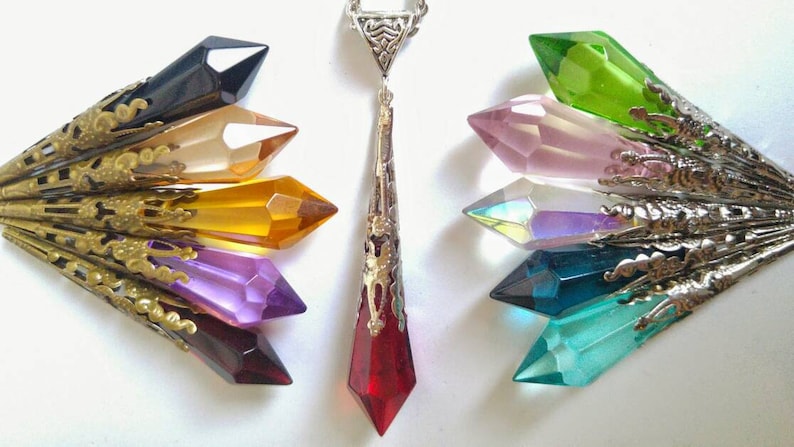May include: A silver chain necklace with a red crystal pendant. The pendant is surrounded by a silver filigree design. The necklace is displayed with seven additional crystal pendants in various colors, including black, orange, yellow, purple, pink, green, and blue.