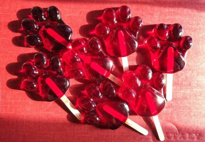 K&ouml;nnte beinhalten: Sechs rote Lutscher in Pfotenform auf Holzstielen. Die Lutscher sind in einem Halbkreis auf einer roten Oberfl&auml;che angeordnet.