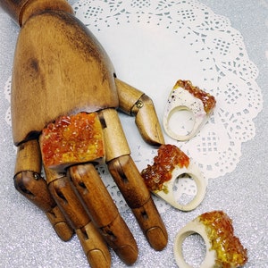 May include: Three resin rings with a white base and orange and yellow resin accents. The rings are displayed on a wooden hand.