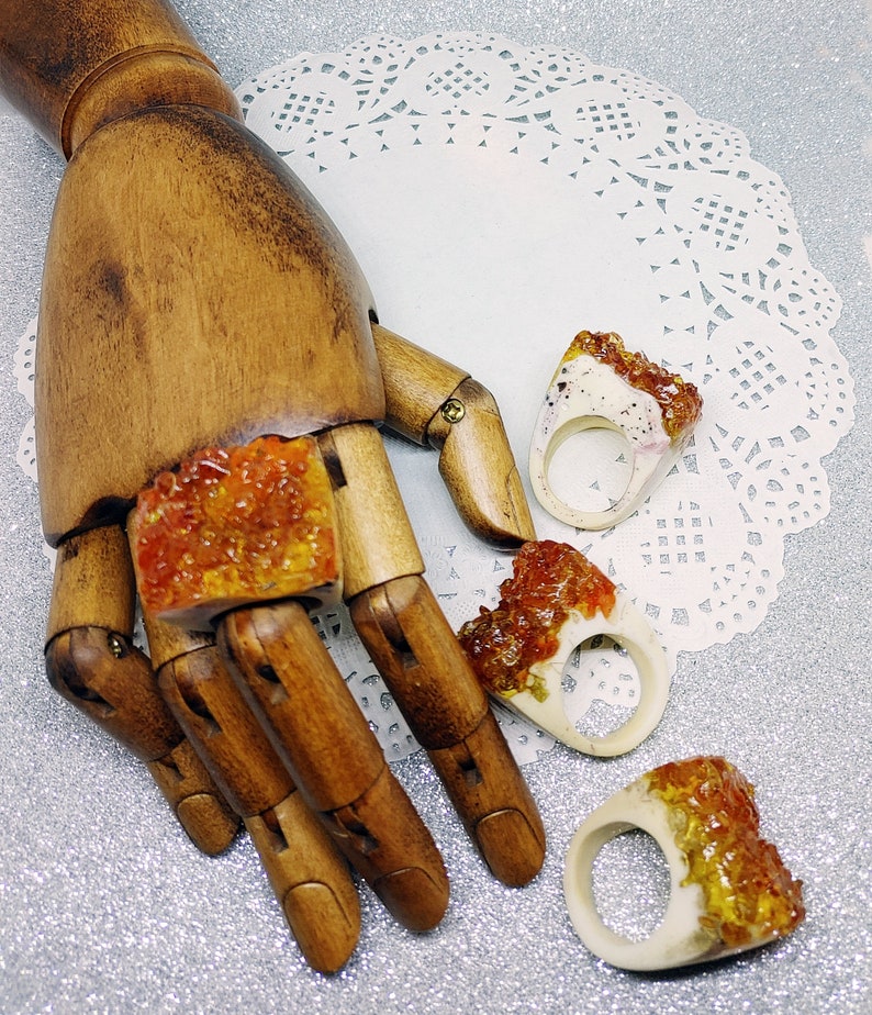 May include: Three resin rings with a white base and orange and yellow resin accents. The rings are displayed on a wooden hand.