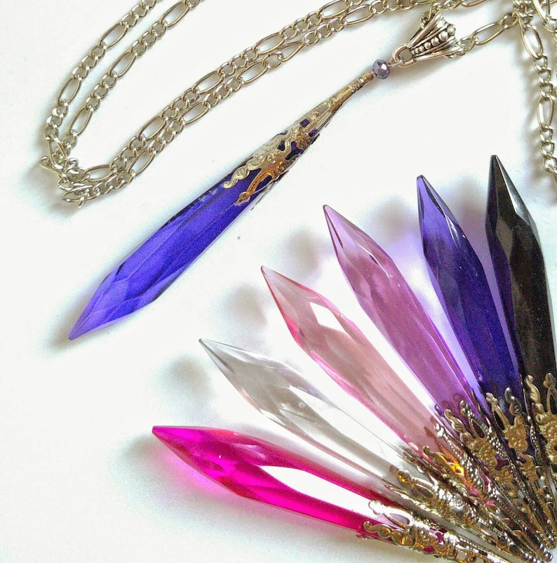 May include: A set of ten crystal pendants in various shades of purple, pink, and clear. The pendants are shaped like long, pointed teardrops and have silver-toned metal accents. The pendants are strung on a silver chain.