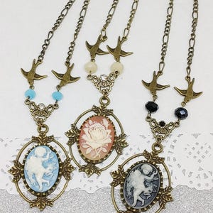 May include: Three gold tone necklaces with cameo pendants. Each necklace features a different cameo design: a rose, a cherub, and a cherub. Each necklace has a chain with two small bird charms.