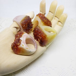 May include: Four resin rings with a translucent white base and a brown, amber-like resin filling. The rings are displayed on a wooden hand.