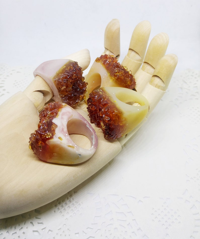 May include: Four resin rings with a translucent white base and a brown, amber-like resin filling. The rings are displayed on a wooden hand.