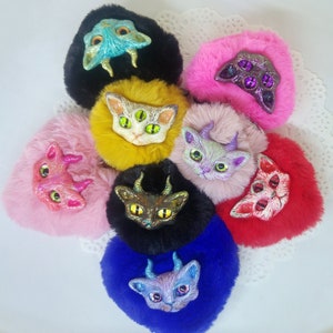 May include: Eight colourful fuzzy pom-poms with different coloured cat faces with horns and eyes. The pom-poms are arranged in a circle on a white surface.