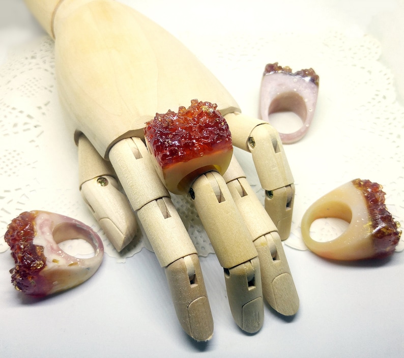 May include: Four resin rings with a crystal-like texture. The rings are in shades of orange, red, and white. The rings are displayed on a wooden hand.