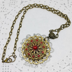 Puede incluir: Un collar de oro y plata con un gran colgante adornado con un diseño de flor roja. El colgante está hecho de metal y tiene un patrón de filigrana. El collar está hecho de un estilo de cadena de eslabones.