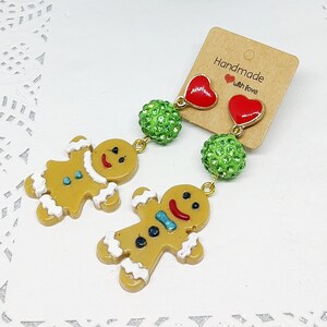 Può includere: Un paio di orecchini a forma di omino di pan di zenzero con dettagli verdi e rossi. Gli orecchini presentano un ciondolo a forma di omino di pan di zenzero, una perla verde scintillante, un ciondolo a forma di cuore rosso e un'altra perla verde scintillante. Gli orecchini sono fatti a mano con amore.