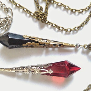 May include: Two silver chains with pendants. The top pendant is a black crystal with gold filigree. The bottom pendant is a red crystal with silver filigree.