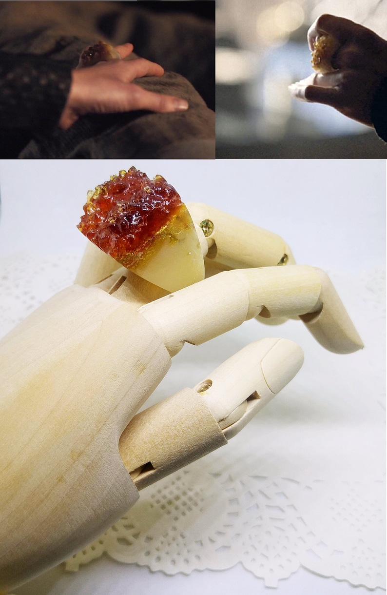 May include: A red and gold resin ring with a unique, crystal-like design. The ring is displayed on a wooden hand model.