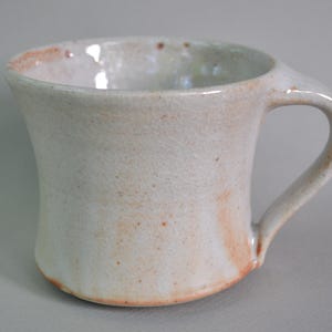 May include: A handmade ceramic mug with a curved handle. The mug has a white glaze with subtle brown and orange accents. The interior and rim show a slightly darker hue. The mug's design is simple and elegant.