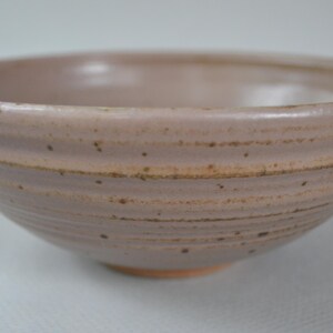 May include: A handmade ceramic bowl in a muted mauve color. The bowl has a textured, ridged exterior and a smooth interior. Small dark speckles dot the surface, adding to its rustic charm. The base is a natural terracotta color.