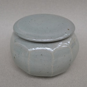 May include: A light blue ceramic jar with a matching lid. The jar has a rounded, faceted body and a smooth, circular lid. The ceramic has a speckled appearance.