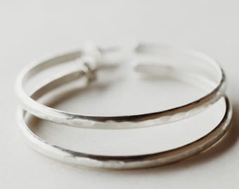 Könnte beinhalten: Ein silberfarbenes Armband mit Doppelband und gehämmertem Muster. Das Armband hat ein schlichtes, minimalistisches Design und ist vor einem neutralen Hintergrund platziert. Die Bänder sind etwa 1,3 cm breit.
