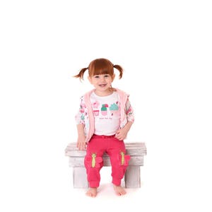 May include: A young girl with red hair sits on a white wooden bench wearing a pink jacket, a white t-shirt with a cupcake and cactus design, and pink pants with butterfly appliques.