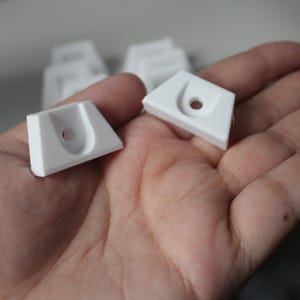 May include: Two white plastic corner brackets with a hole in the centre. The brackets are designed to be used for mounting shelves or other objects.