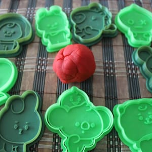 Puede incluir: Nueve cortadores de galletas de plástico verde con forma de personajes de dibujos animados, incluyendo un corazón, un conejito y un oso. Una bola de plastilina roja se encuentra en el centro de los cortadores.