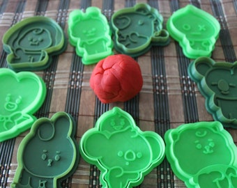 May include: Nine green plastic cookie cutters shaped like cartoon characters, including a heart, a bunny, and a bear. A ball of red playdough is in the center of the cutters.