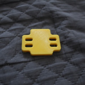 May include: A yellow, cross-shaped plastic component with four rectangular holes. The object is centered on a dark gray, quilted fabric background. The component appears to be 3D-printed.
