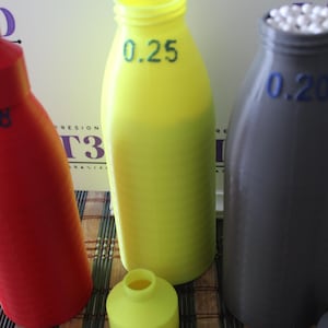 Pode incluir: Três garrafas impressas em 3D em vermelho, amarelo e cinza. A garrafa vermelha tem o número 0,28, a amarela 0,25 e a cinza 0,20 impressos. A garrafa cinza é preenchida com contas brancas. As garrafas estão em uma superfície listrada.