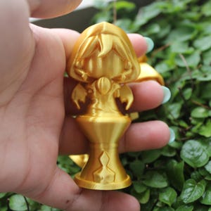 Puede incluir: Una figurita dorada de una persona con una capa con capucha y de pie sobre un pedestal. La figurita est&aacute; detallada con una cara, manos y una capa fluida.