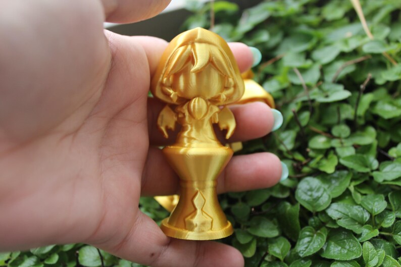 Puede incluir: Una figurita dorada de una persona con una capa con capucha y de pie sobre un pedestal. La figurita est&aacute; detallada con una cara, manos y una capa fluida.