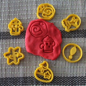 Puede incluir: Un conjunto de cortadores de galletas de plástico amarillo con varios diseños, que incluyen una boca de incendios, una estrella, un remolino, una bolsa de dinero, una calavera y un signo de exclamación. Los cortadores están dispuestos en una estera tejida marrón y negra con una bola de plastilina roja en el centro.
