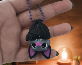 May include: A gray and black bat pendant with purple accents, hanging from a purple beaded chain. The bat has large eyes, a pink nose, and purple-lined ears. The pendant is held in a hand against a blurred background.