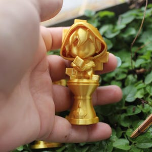 Puede incluir: Una figurita de un personaje con una capa con capucha y un libro en la mano, impresa en 3D en color dorado. La figurita est&aacute; de pie sobre un pedestal.