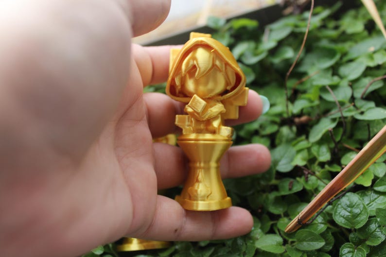 Puede incluir: Una figurita de un personaje con una capa con capucha y un libro en la mano, impresa en 3D en color dorado. La figurita est&aacute; de pie sobre un pedestal.