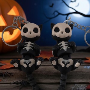 Peut inclure: Deux porte-clés chat squelette noirs et blancs avec une citrouille et un décor d'Halloween en arrière-plan. Les porte-clés ont un motif de squelette sur le corps et la tête. La citrouille est sculptée avec un visage de lanterne.