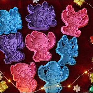 May include: A collection of colorful Stitch-themed cookie cutters in various Christmas designs. The cutters are in shades of blue, pink, and purple, arranged on a red surface with festive decorations, including lights and greenery.