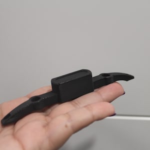 Può includere: Un oggetto in plastica nera stampato in 3D con due bracci e una base rettangolare. L'oggetto è progettato per tenere qualcosa in posizione.