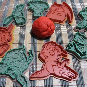 Puede incluir: Un conjunto de ocho cortadores de galletas con varios personajes de dibujos animados. Los cortadores están hechos de plástico verde y rojo y están dispuestos en una estera de bambú tejida. Una bola de plastilina roja se encuentra en el centro de la disposición.