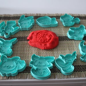 May include: A set of 12 teal blue plastic cookie cutters featuring various cartoon characters and a fish shape. A red playdough fish is in the center of the circle of cutters.
