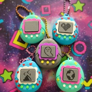 Puede incluir: Cinco llaveros digitales de mascotas de estilo retro. Cada llavero presenta una pantalla que muestra un gráfico pixelado, con diseños que incluyen un corazón, un fantasma y un mundo. Los llaveros son en tonos de azul, verde y gris, con acentos rosas y blancos.