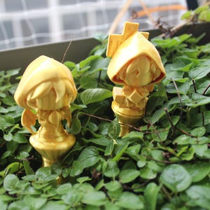 Puede incluir: Dos figuritas doradas de personas con capuchas de pie en un lecho de plantas verdes.