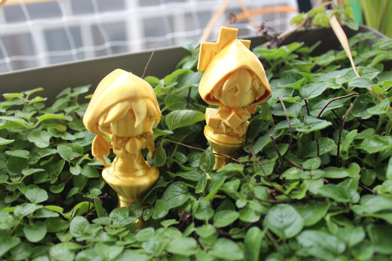 Puede incluir: Dos figuritas doradas de personas con capuchas de pie en un lecho de plantas verdes.