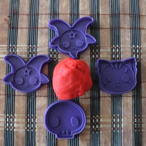 Puede incluir: Cinco cortadores de galletas de plástico morado sobre una estera de bambú marrón. Los cortadores tienen forma de calavera, gato y dos cabras. Hay una bola de plastilina roja en el centro.