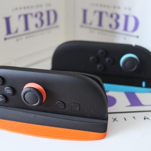 Peut inclure: Deux manettes de jeu noires et oranges avec des boutons et des joysticks. Une manette a un joystick noir avec un anneau orange. L'autre manette est noire avec un accent bleu. Le texte "LT3D" est visible en arrière-plan.