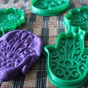 Puede incluir: Cuatro cortadores de galletas de plástico verde con diseños intrincados, incluyendo una mano de hamsa, un mandala y un círculo. Un cortador de galletas de mano de hamsa se está utilizando para presionar una plastilina morada.