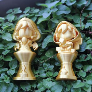 Puede incluir: Dos figuritas doradas de personas con capas con capucha se colocan sobre bases decorativas. Las figuritas est&aacute;n sobre un lecho de hojas verdes.