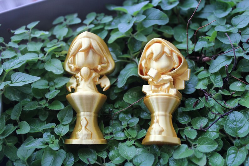 Puede incluir: Dos figuritas doradas de personas con capas con capucha se colocan sobre bases decorativas. Las figuritas est&aacute;n sobre un lecho de hojas verdes.