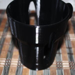 Könnte beinhalten: Ein schwarzer Plastik-Becherhalter mit einer strukturierten Oberfläche. Der Becherhalter hat einen Griff an der Seite.