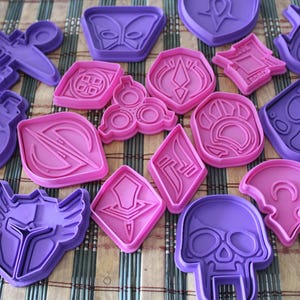 May include: An assortment of purple and pink plastic cookie cutters in various shapes, including a skull, paw print, and geometric designs. The cutters are arranged on a striped surface.