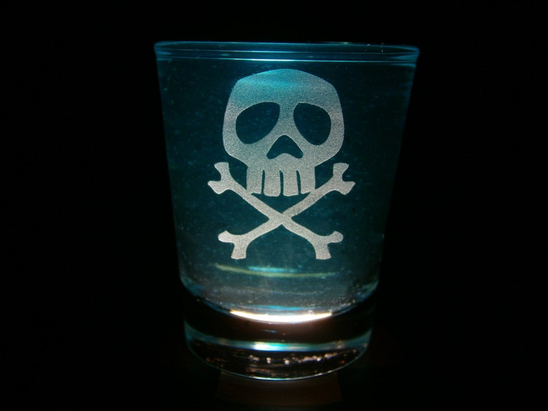 Puede incluir: Un vaso de chupito de vidrio transparente con un dise&ntilde;o de calavera y huesos cruzados grabado en el lateral. El vaso est&aacute; lleno de un l&iacute;quido azul.